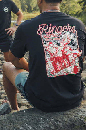 A person sits on a rocky riverside wearing a Ringers Western Whiskey Business Loose Fit T-Shirt, featuring a large red and white back graphic with a cowboy riding a bucking horse and the text βRingers Western Whiskey Business.β.