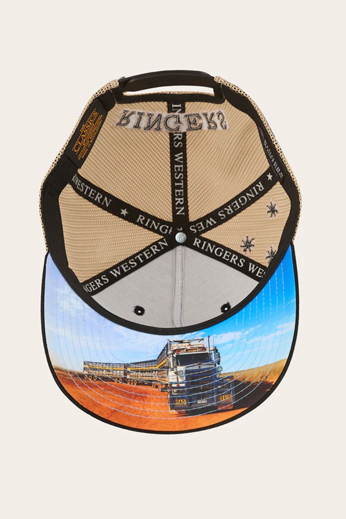 The Ringers Western Workhorse Trucker Cap features "Ringers Western" branding inside and a brim printed with a large truck on a red dirt road beneath a blue sky. It comes with an adjustable snapback for the perfect fit.