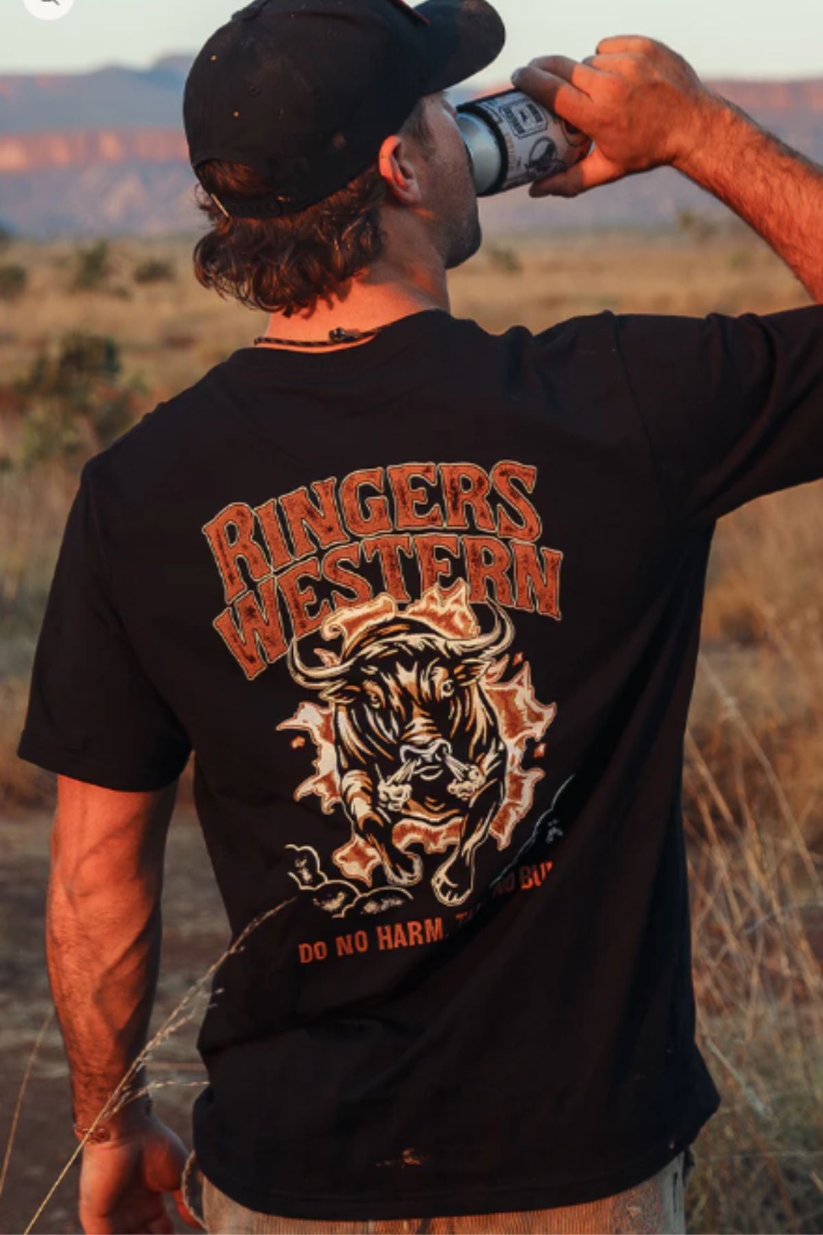 A man in a black Ringers Western Do No Harm Take No Bull Loose Fit T-Shirt drinks from a can in a dry outdoor landscape.