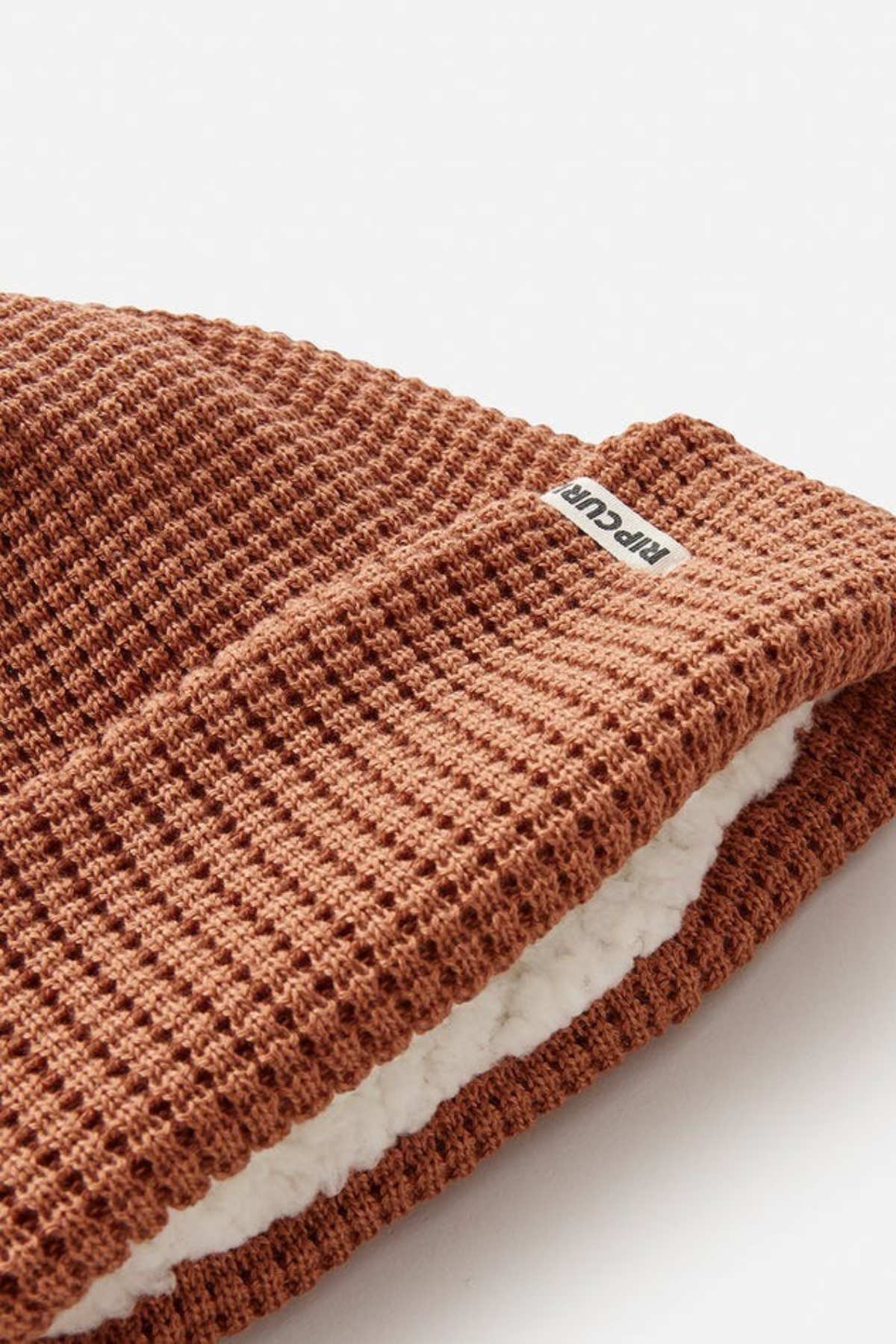 A close-up of the Rip Curl Anoeta Reg Pom Pom Beanie, a brown knitted winter hat with a waffle pattern and teddy fur lining made from recycled polyester. A small label in the seam suggests its cozy origin.
