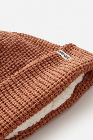 A close-up of the Rip Curl Anoeta Reg Pom Pom Beanie, a brown knitted winter hat with a waffle pattern and teddy fur lining made from recycled polyester. A small label in the seam suggests its cozy origin.