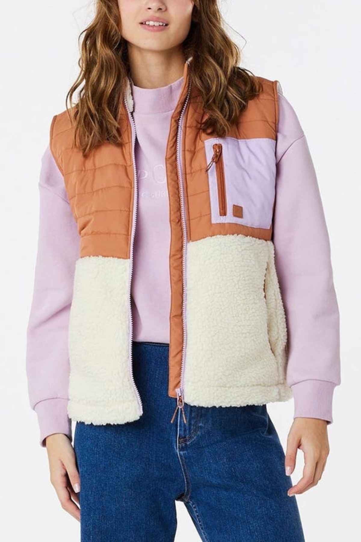 A person with long brown hair is wearing a Rip Curl Anti-Series Anoeta Fleece Vest with brown, light purple, and white sections over a light purple sweatshirt and blue jeans. The vest has a zip chest pocket, and they are standing against a plain white background.