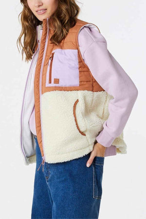 A person sports a color-blocked Rip Curl Anti-Series Anoeta Fleece Vest featuring orange, light purple, and white panels over a lavender hoodie. Their wavy hair pairs well with blue jeans, and the shower resistance keeps them dry in style.