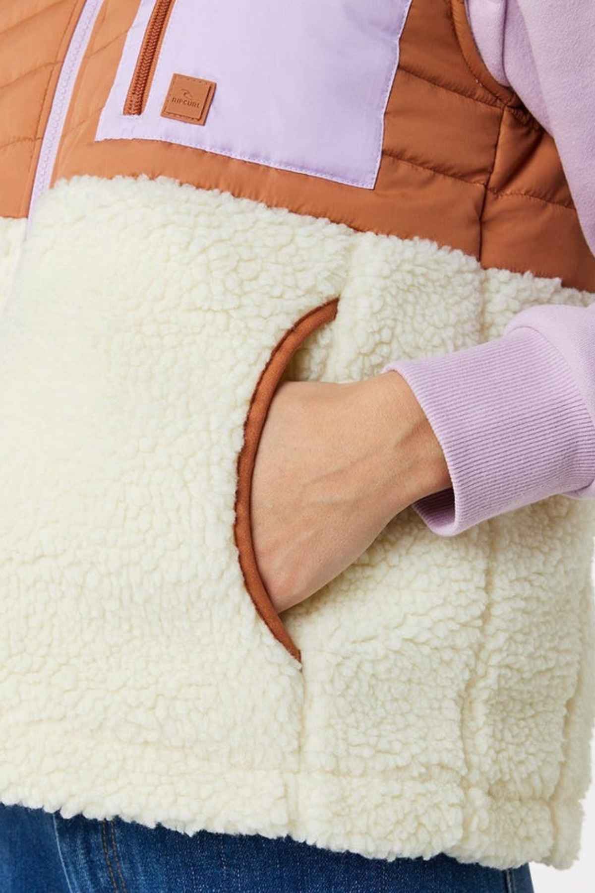 A close-up of a hand in the pocket of the Rip Curl Anti-Series Anoeta Fleece Vest shows off its color-blocked design with a shower-resistant brown top, lilac middle, and cream-colored sherpa bottom. Perfect for pairing with denim jeans, it blends style and practicality effortlessly.