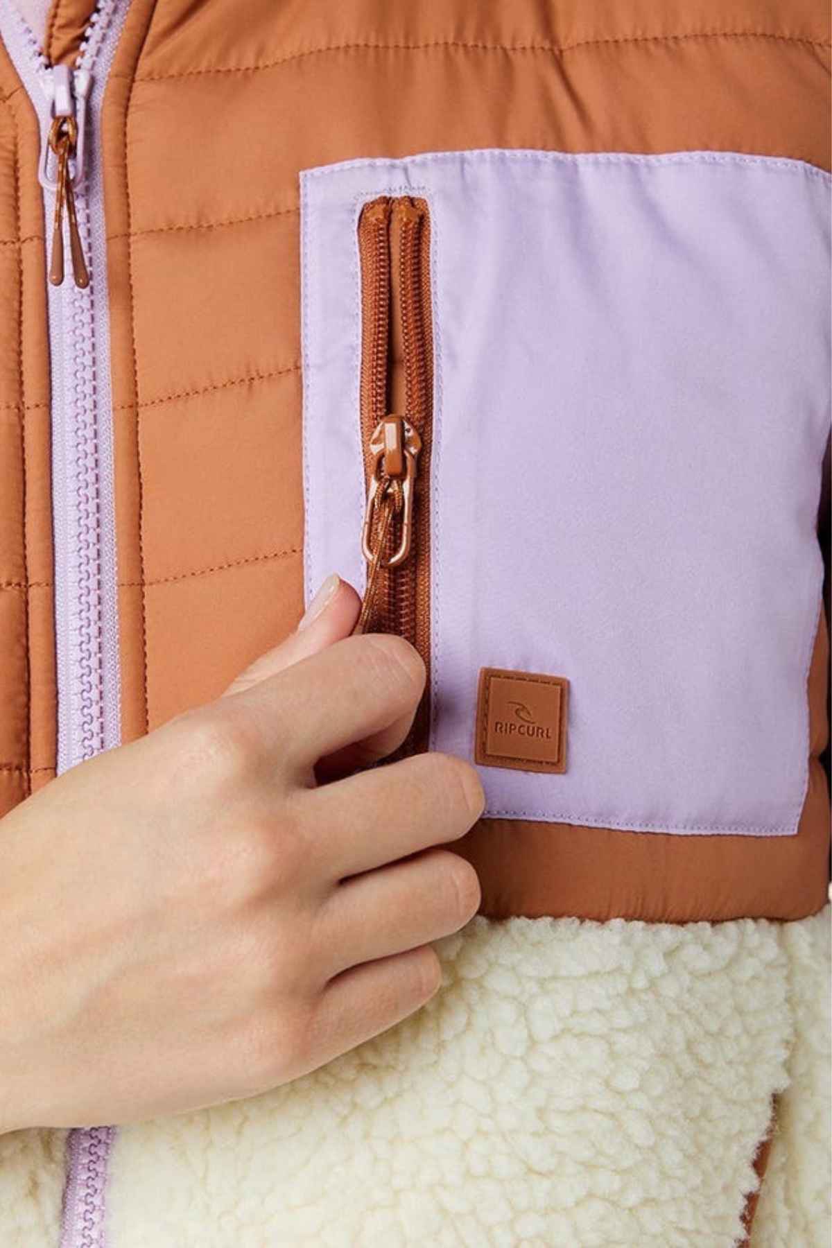 Close-up of someone wearing a Rip Curl Anti-Series Anoeta Fleece Vest in brown and light purple with shower resistance. It features a zip-up pocket with a leather logo and fluffy white texture on the lower part, offering both style and practicality.