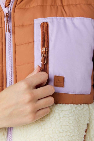 Close-up of someone wearing a Rip Curl Anti-Series Anoeta Fleece Vest in brown and light purple with shower resistance. It features a zip-up pocket with a leather logo and fluffy white texture on the lower part, offering both style and practicality.