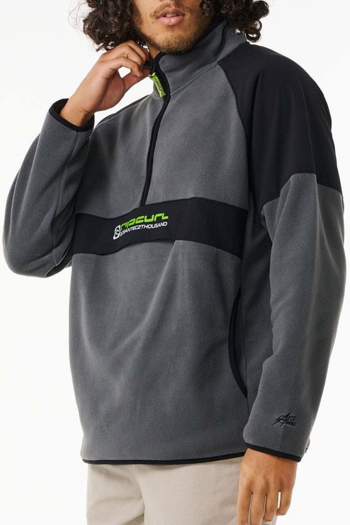 A person wears the Rip Curl Anti Series Archive Zip Crew, a gray and black fleece pullover with a half-zip and chest logo, holding the collar with one hand. The background is plain white.