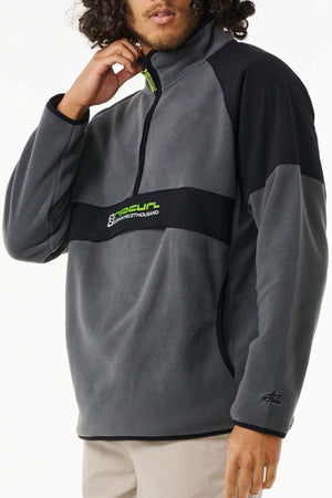 A person wears the Rip Curl Anti Series Archive Zip Crew, a gray and black fleece pullover with a half-zip and chest logo, holding the collar with one hand. The background is plain white.