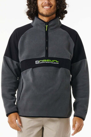 A person wears the Rip Curl Anti Series Archive Zip Crew, a gray and black fleece pullover with water repellent finish, front zipper, and green logo text, standing against a plain white background.