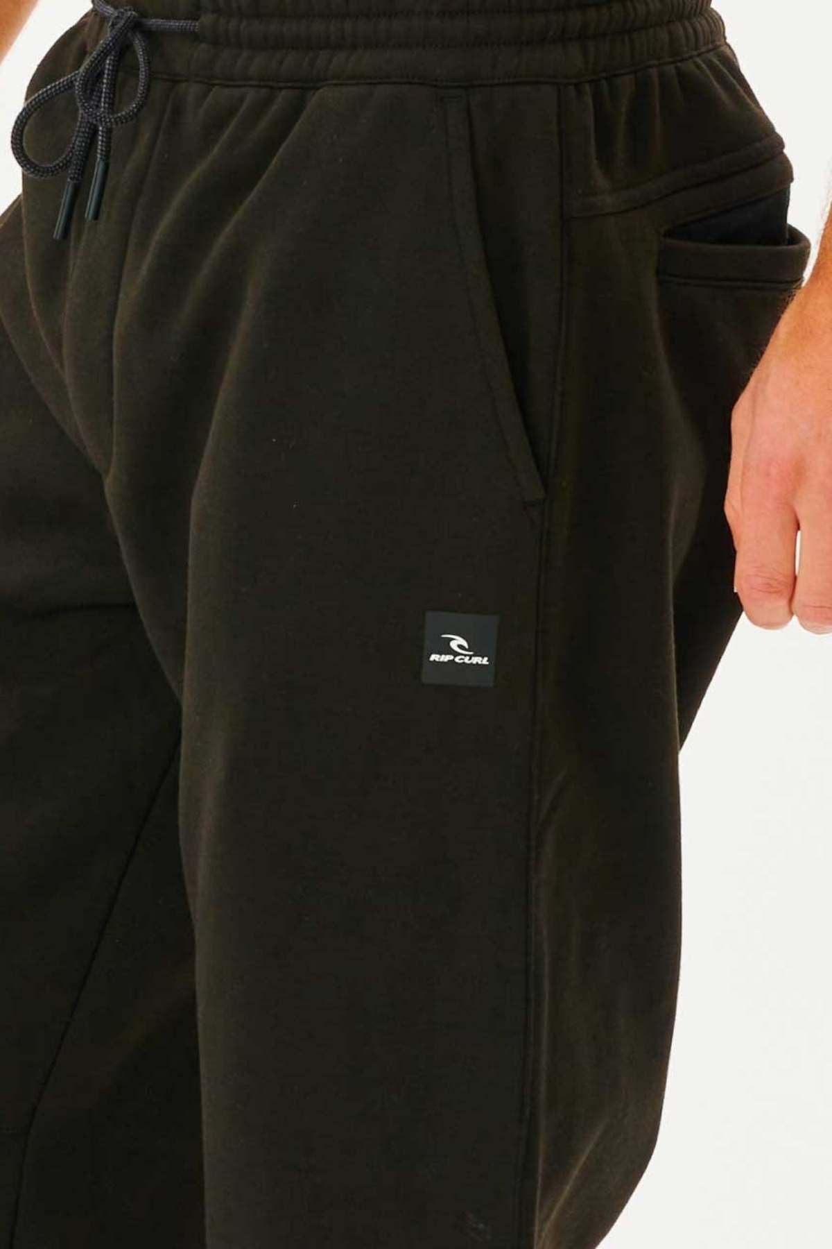 Close-up of a person wearing Rip Curl Departed Anti Series Trackpant, featuring a drawstring waist, side pockets, bonded polar fleece fabric, and a small Rip Curl logo patch on the left thigh. The personโs right hand is near the pocket.