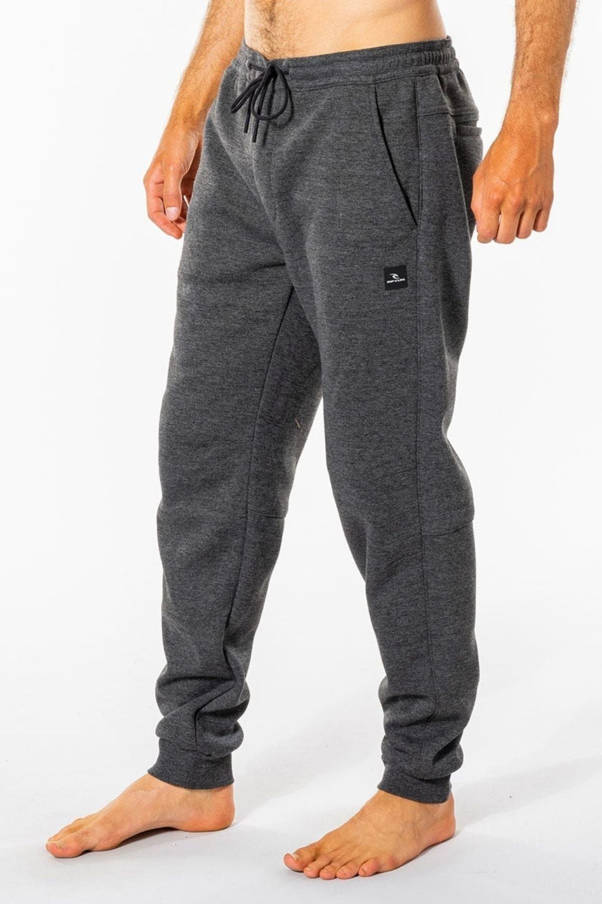 The person stands barefoot in Rip Curl Departed Anti Series Trackpants, dark gray with an elastic waistband and drawstring. These water-repellent pants have side pockets, a small black logo on the left thigh, and are set against a plain white background.