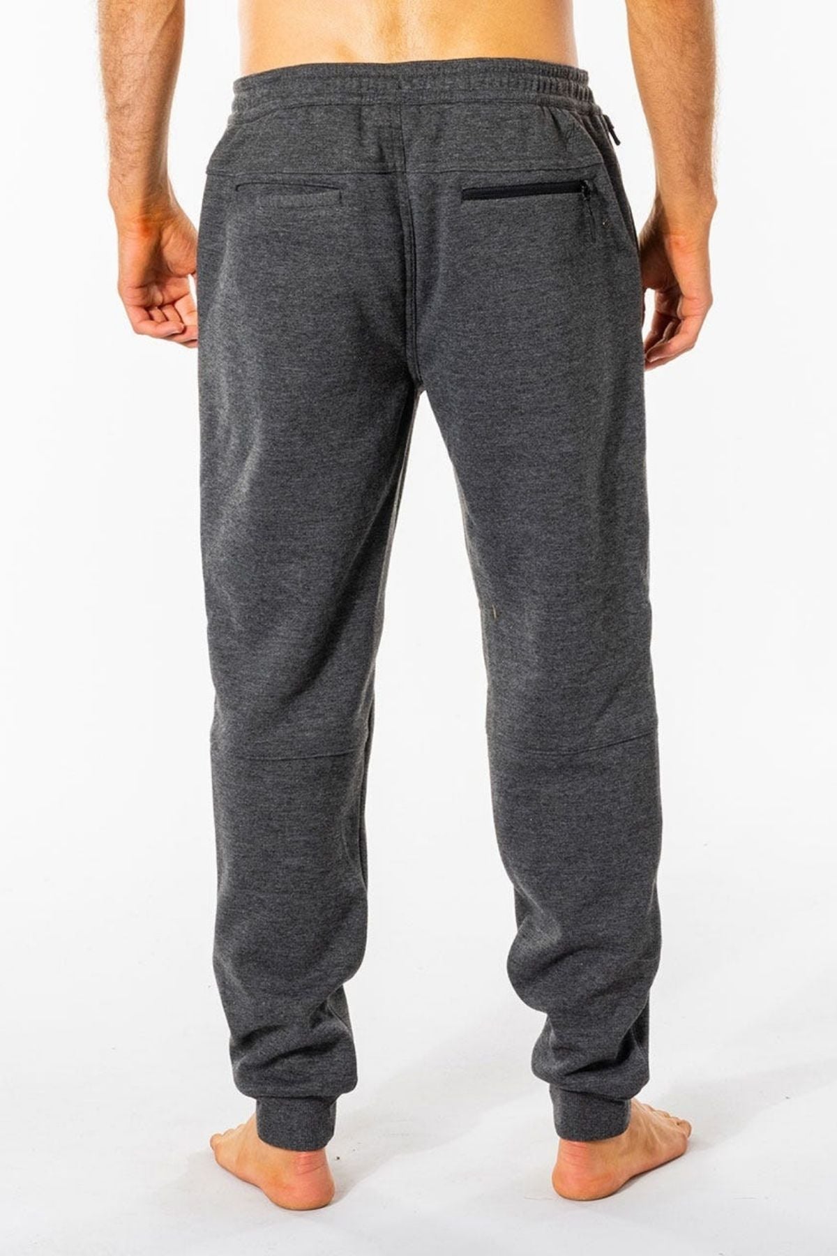 A person stands barefoot on a light surface, facing away, wearing Rip Curl Departed Anti Series Trackpant. These dark gray joggers feature an elastic waistband and back pockets, crafted from bonded polar fleece. The clean white backdrop highlights their sleek design.