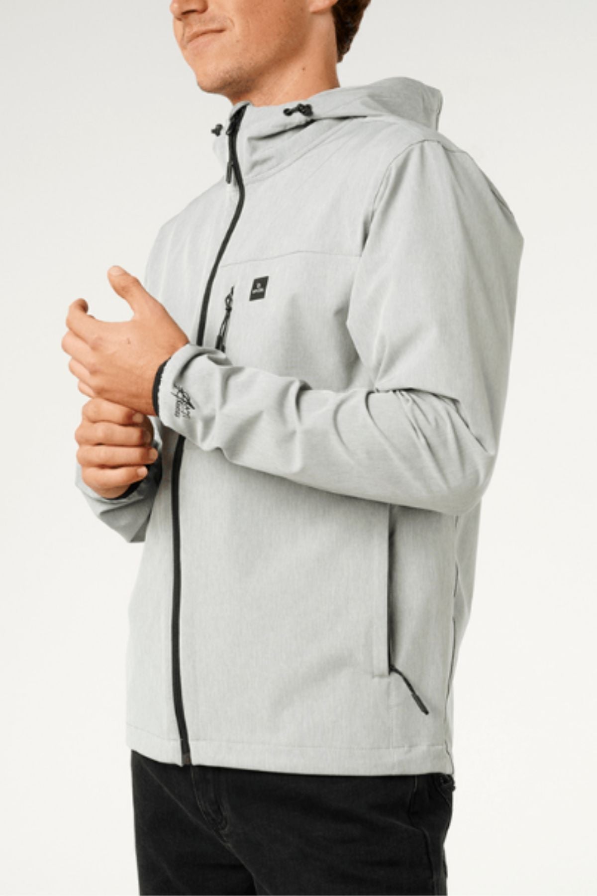 A person wears the Rip Curl Anti Series Elite Jacket in light gray stretch polyester with black zippers, paired with black pants, standing against a plain background. Their face is partially visible and hands are clasped together.