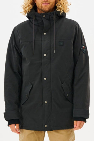 A person with long curly hair wears a black Rip Curl Anti Series Exit Jacket with snap buttons, front pockets, and a sleeve patch, paired with tan pants. The durable water-resistant jacket stands out against a plain background.