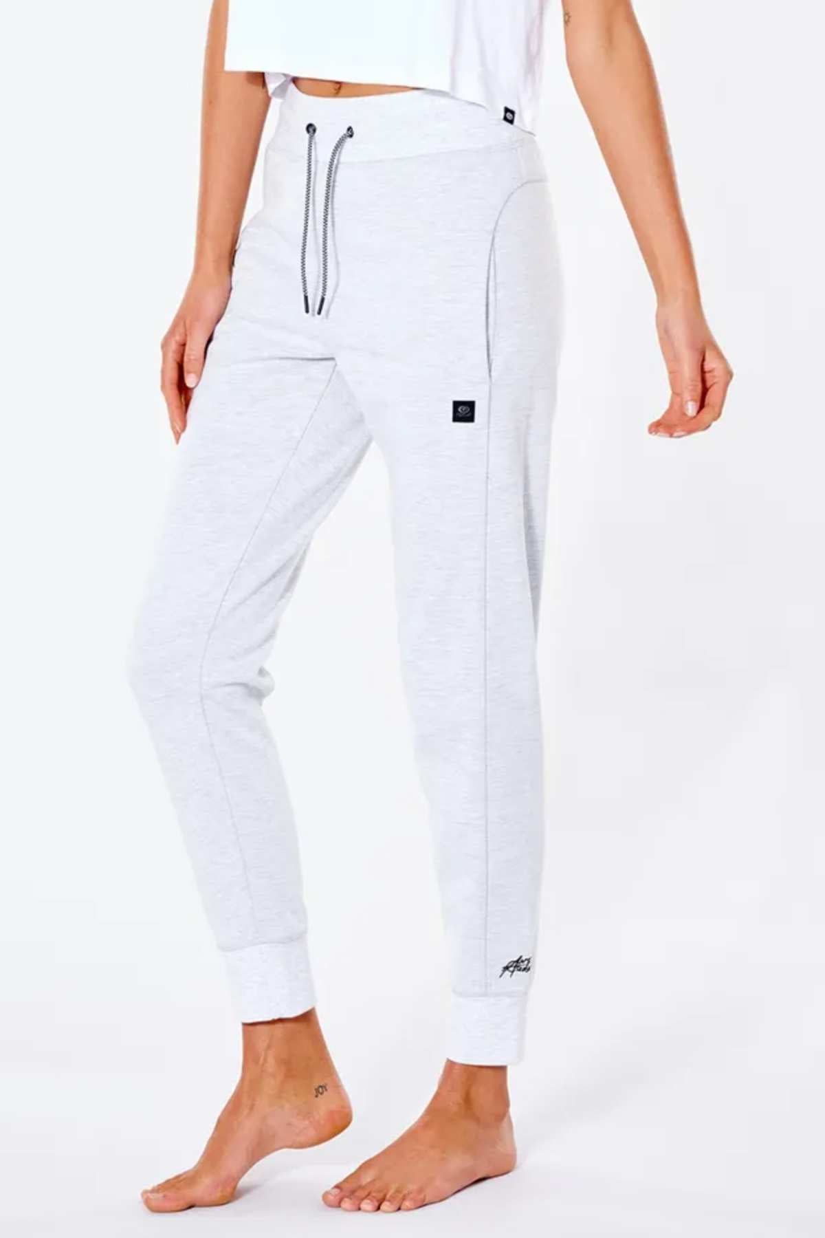 A barefoot person is wearing light gray Rip Curl Anti Series Flux II Trackpant with black stitching, drawstring waist, thermal protection, and cuffed ankles. The trackpants feature a small logo near the pocket. The upper body in a white top is partially visible.