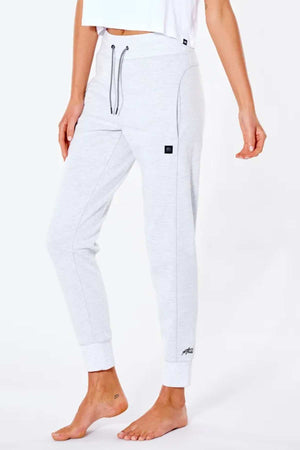 A barefoot person is wearing light gray Rip Curl Anti Series Flux II Trackpant with black stitching, drawstring waist, thermal protection, and cuffed ankles. The trackpants feature a small logo near the pocket. The upper body in a white top is partially visible.
