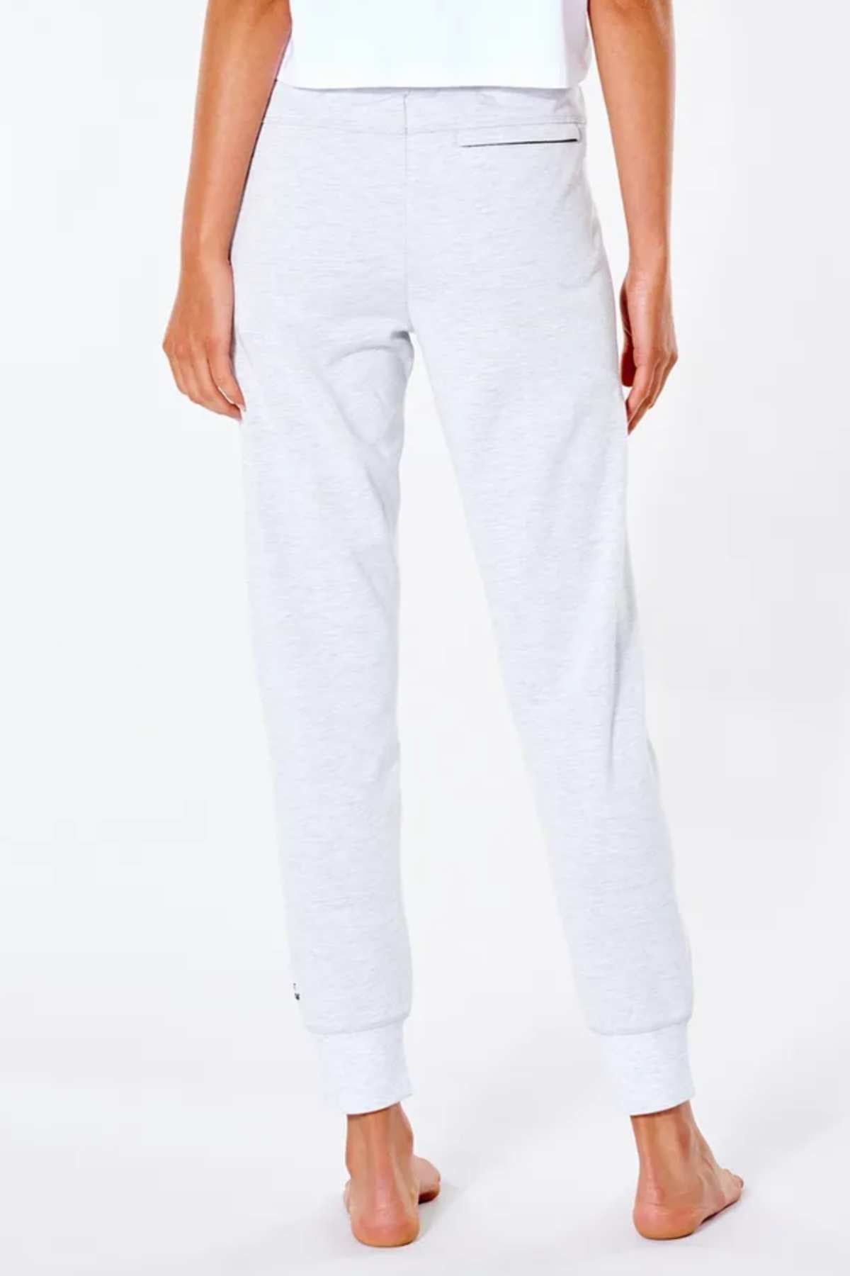 On a white backdrop, a person stands barefoot wearing Rip Curl Anti Series Flux II Trackpant in light gray and a white top, displaying the lower body from shoulders to feet.