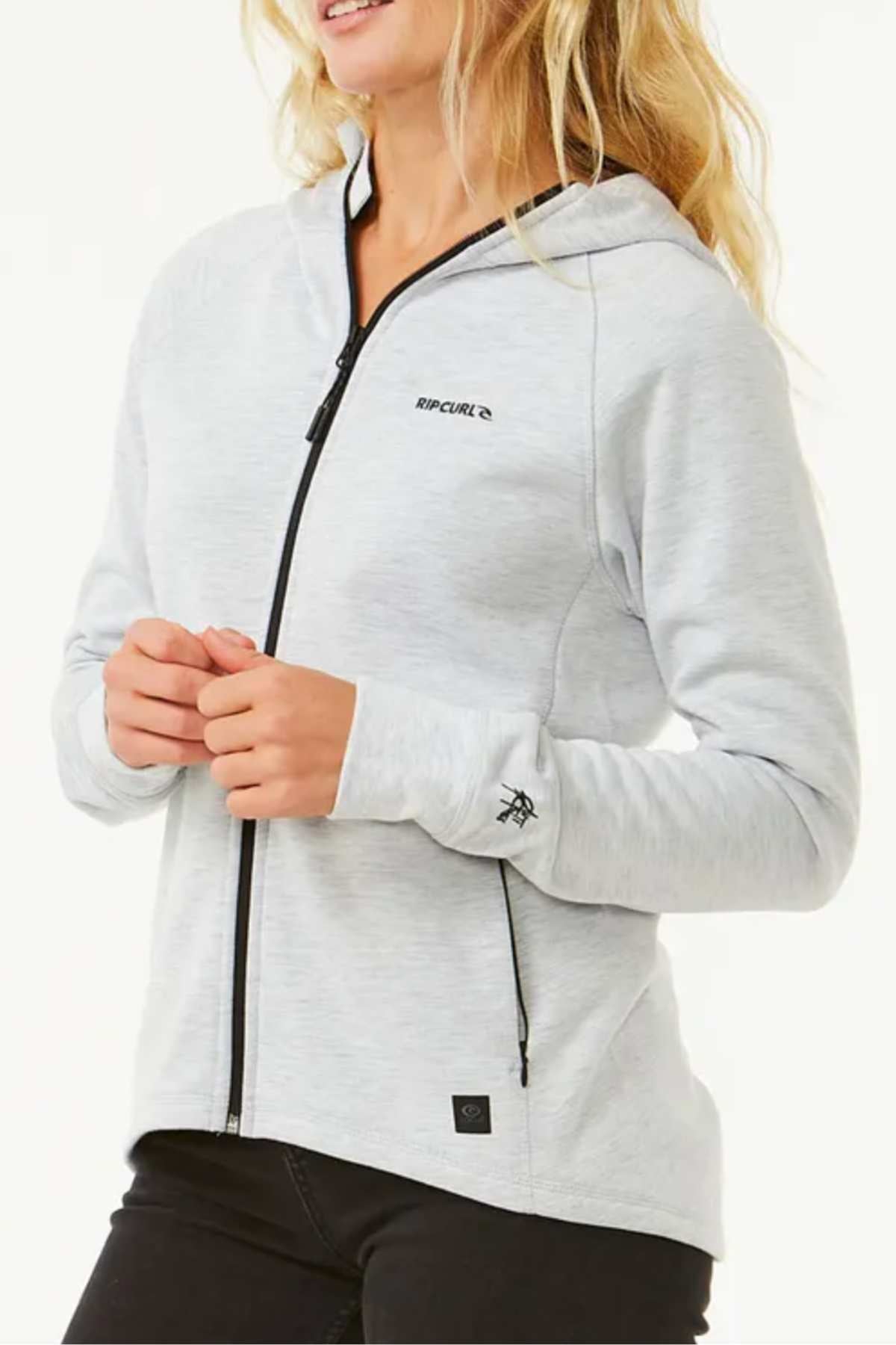 A woman stands against a white background wearing the Rip Curl Anti Series Flux III Zip light gray hoodie with black trim and black pants, featuring bonded polar fleece for warmth.
