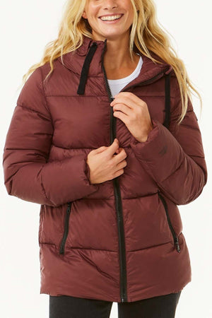 A person with long, light-colored hair is smiling and wearing a maroon puffer jacket from the Rip Curl Anti Series Insulated Jacket collection. The jacket features a quilted design, black zippers, and drawstrings. They are partially unzipping it against a plain, light-colored background.