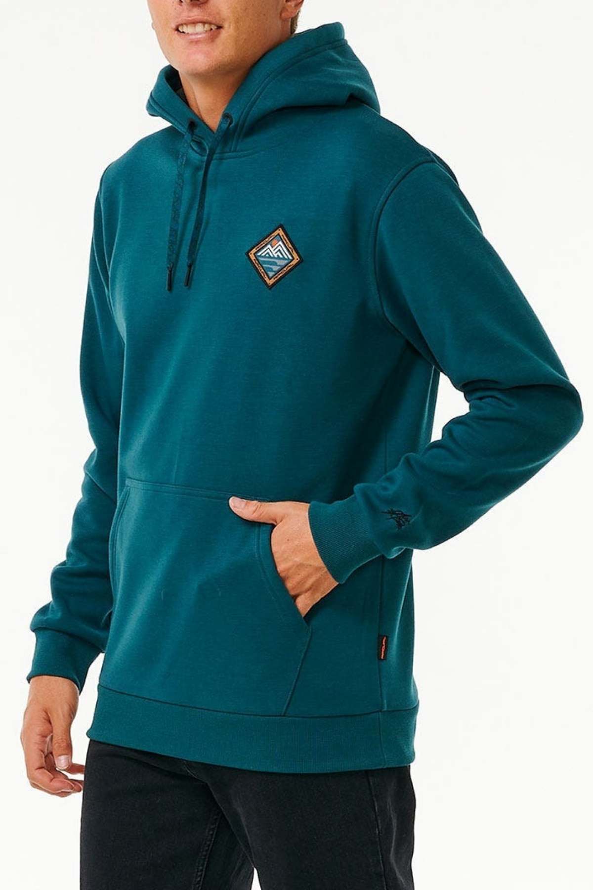 Against a white backdrop, a person models the Rip Curl Anti Series Journey Hood—a teal hoodie with a geometric patch and thumb holes. One hand rests in the pocket, showcasing the drawstrings. Paired with black pants, this water-repellent outfit merges style and function effortlessly.