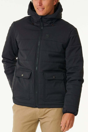 A person is wearing a dark Rip Curl Anti Series Ridge Jacket, featuring a front zipper and snap-button pockets, perfect for the outdoor enthusiast. The jacket is stylishly paired with beige pants. The background is plain and light-colored, while the person's face is partially visible, looking to the side.