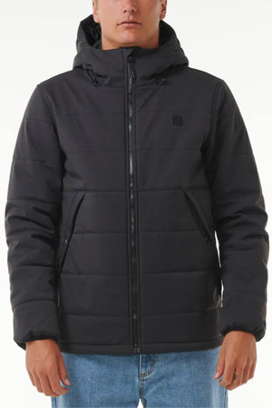 Wearing a black Rip Curl Anti Series Ridge Jacket with a hood, quilted padded lining, and two side pockets, styled with light blue jeans. The jacket’s relaxed fit contrasts with the plain light background.