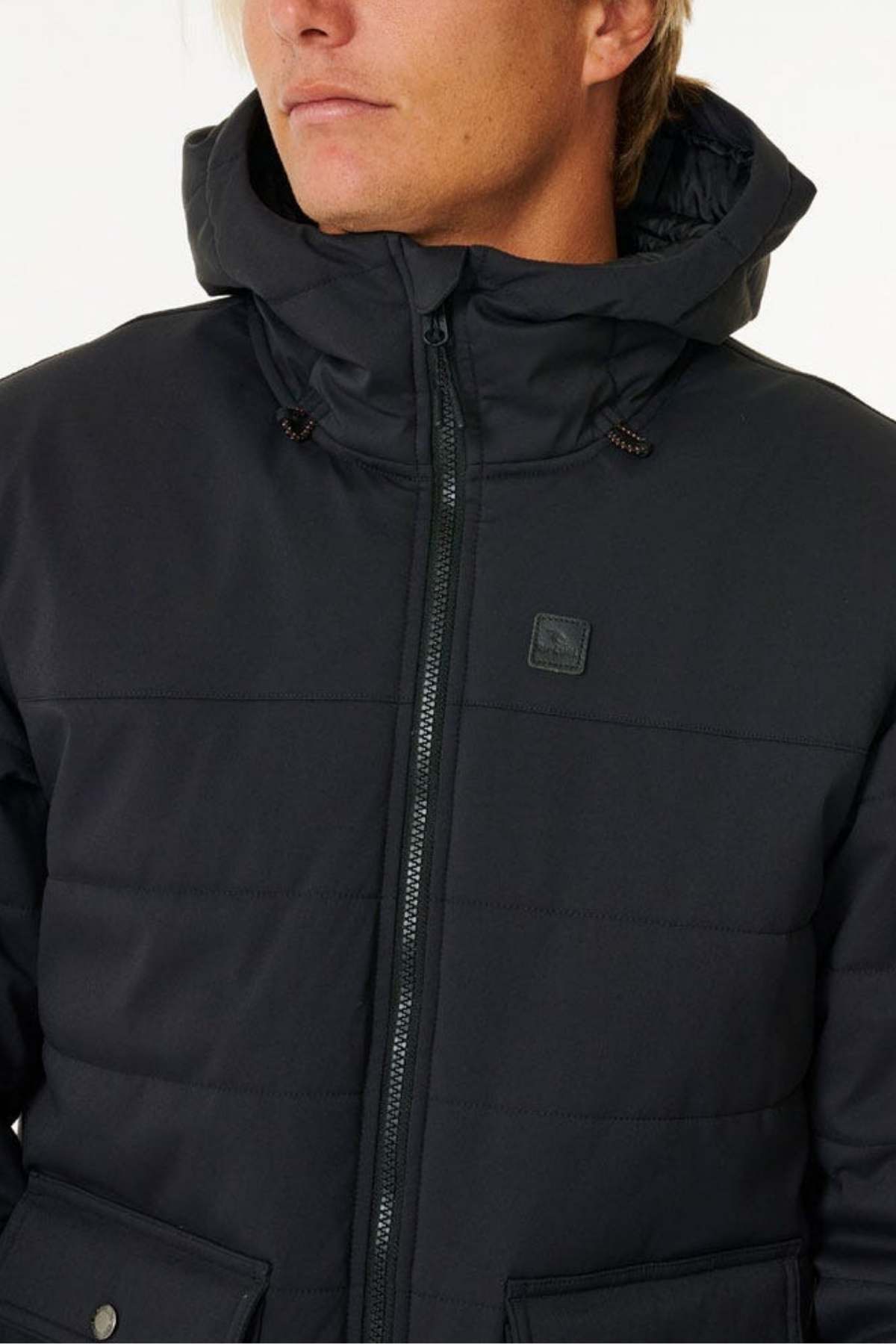 A person wears a dark-colored Rip Curl Anti Series Ridge Jacket with the hood up and the jacket zipped. The jacket features a front zipper, a logo on the chest, and noticeable stitching across the chest. Its durable water repellent (DWR) finish makes it perfect for any outdoor enthusiast. The person is visible from the shoulders up against a plain background.