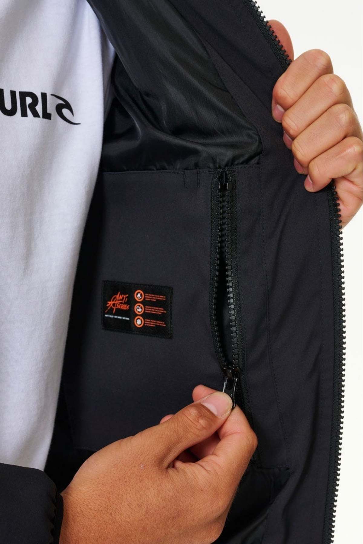A person unzips the pocket of a Rip Curl Anti Series Ridge Jacket, showcasing its black label with orange text. The outdoor enthusiast, dressed in a white shirt with partially visible black lettering, holds the jacket open with one hand while using the other to unzip the pocket.