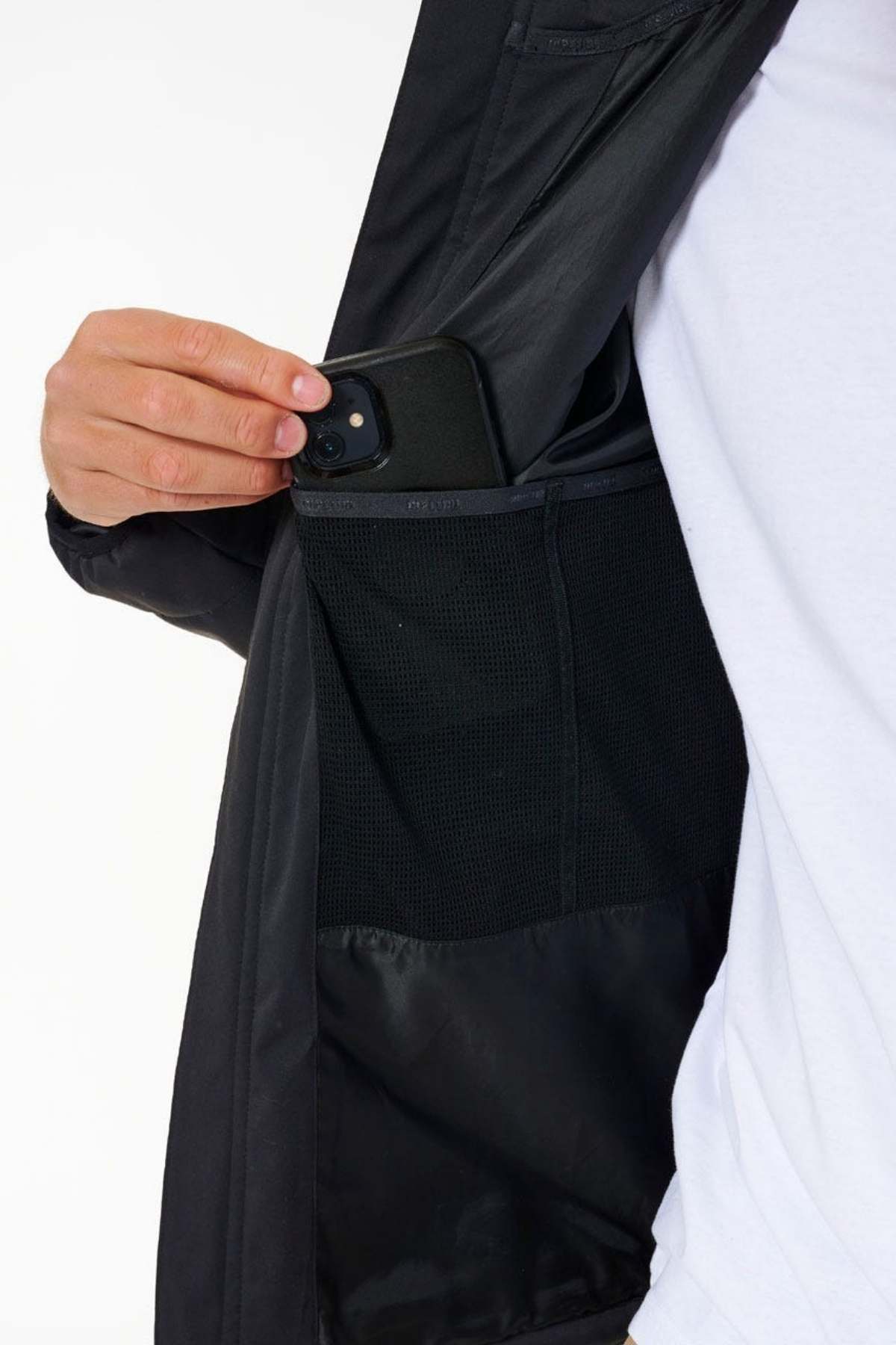 An outdoor enthusiast, dressed in a white shirt and a Rip Curl Anti Series Ridge Jacket, is tucking a black smartphone into the zippered inner pocket. The partially open jacket reveals its inner mesh and fabric compartments lining, ideal for keeping essentials secure and dry thanks to its DWR water repellent feature.