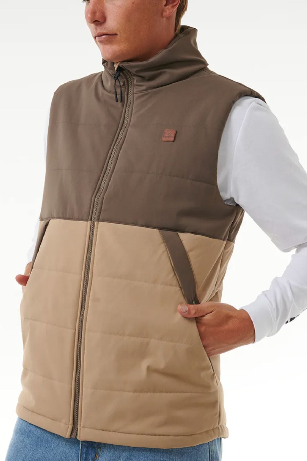 Someone wears the Rip Curl Anti Series Ridge Vest in brown and beige, featuring color-blocking and quilted padded lining, over a white long-sleeve shirt, with hands in the vest pockets; image shows from chest to waist.