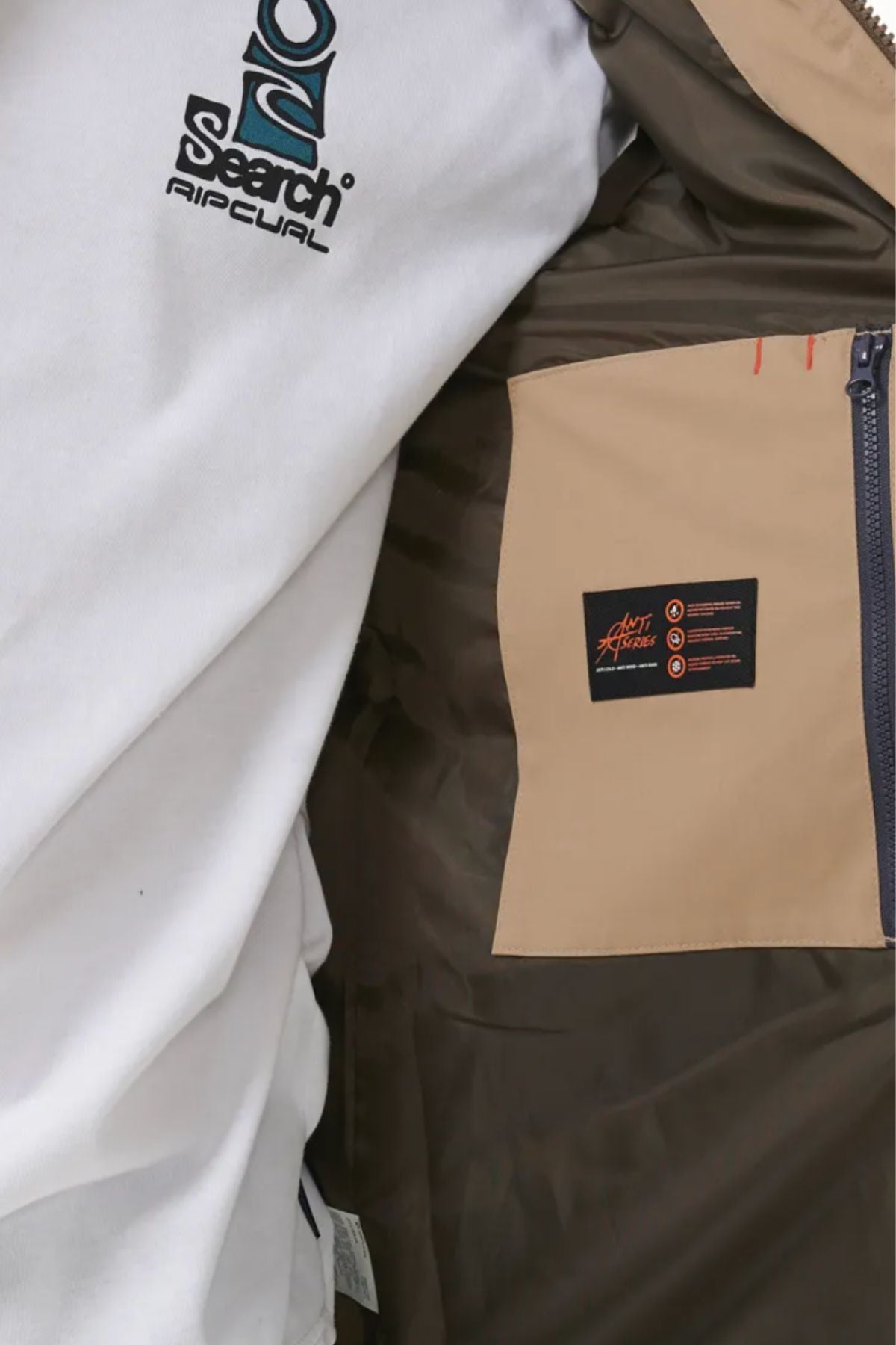 Close-up of someone wearing the Rip Curl Anti Series Ridge Vest, featuring a white shirt, brown jacket with quilted lining, beige pocket, blue zipper, and black "Heat Seeker" label—highlighting its premium comfort.