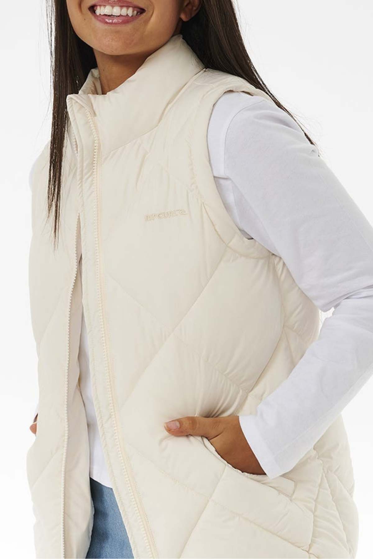 A person wearing a **Rip Curl Anti Series Shorebreak Vest** over a long-sleeve white shirt smiles while posing. They have one hand in their pants pocket and the other holding the edge of the water-repellent vest, with a plain white background behind them.