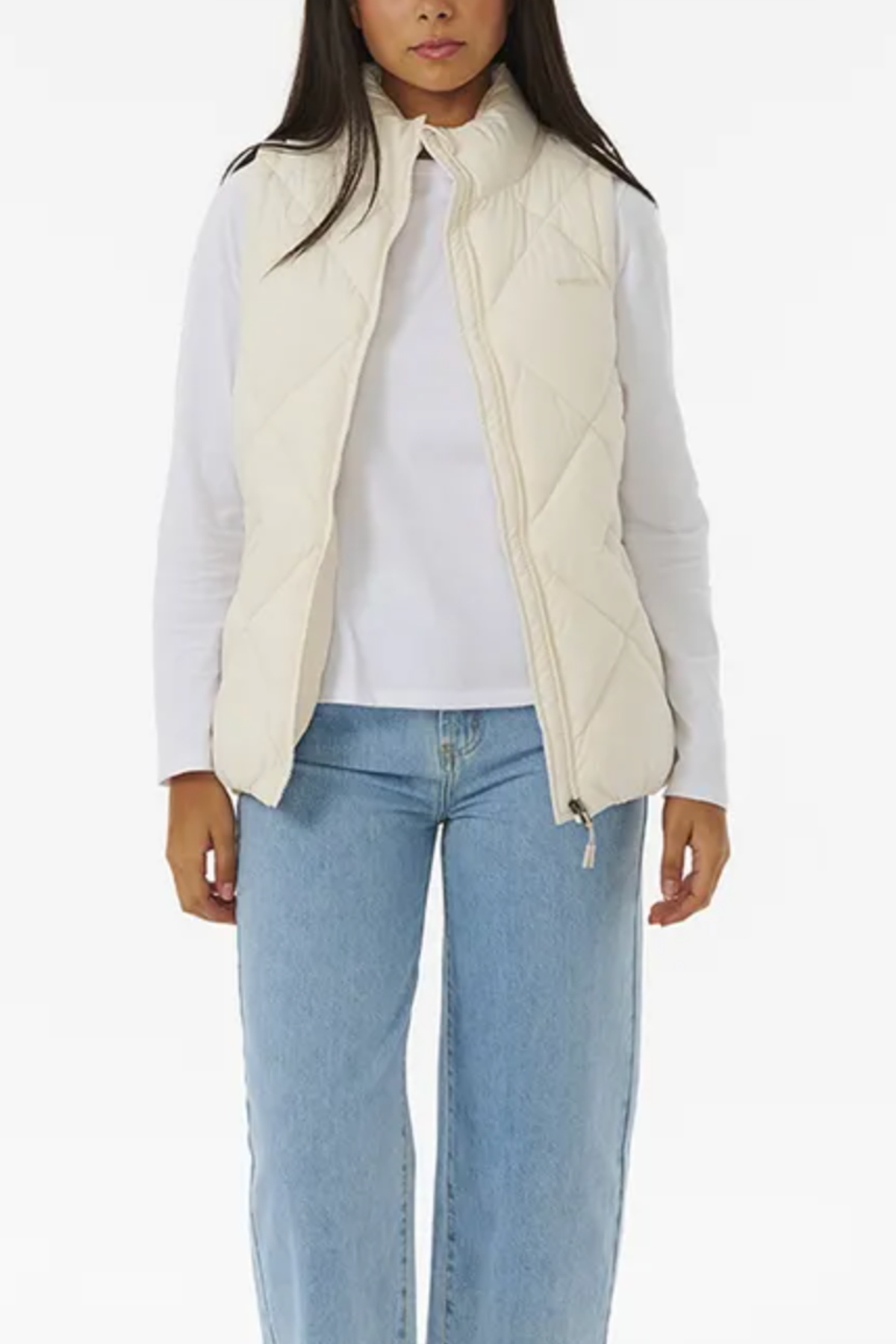 A person is wearing a Rip Curl Anti Series Shorebreak Vest in cream over a white long-sleeved shirt and blue high-waisted jeans. The vest is unzipped, showcasing the shirt beneath. The image features a plain white background.