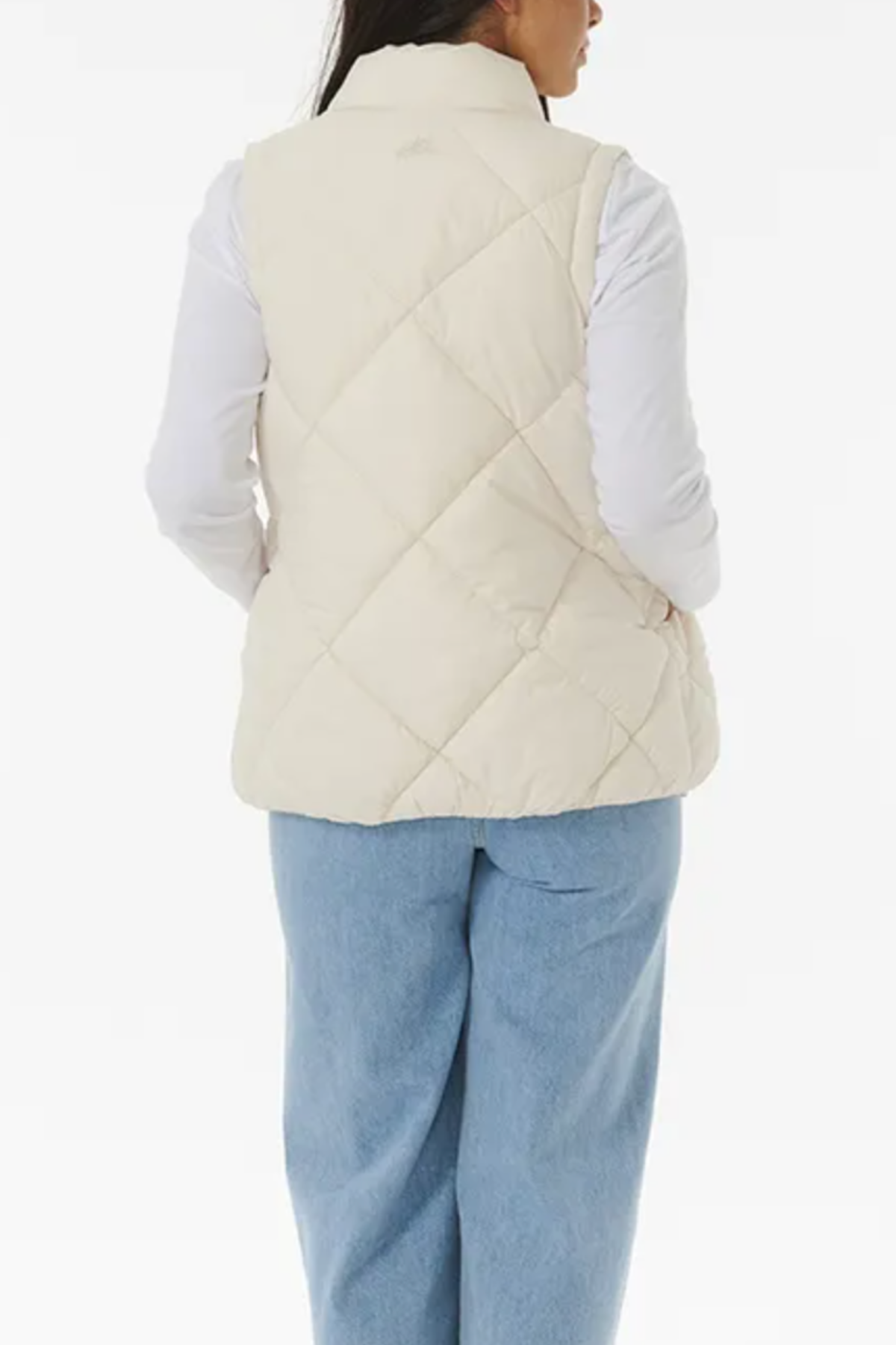 A person is shown from the back, wearing a Rip Curl Anti Series Shorebreak Vest in a cream-colored quilted design over a long-sleeved white shirt and light blue jeans. The background is plain white.