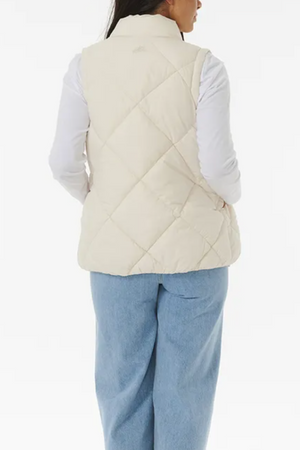 A person is shown from the back, wearing a Rip Curl Anti Series Shorebreak Vest in a cream-colored quilted design over a long-sleeved white shirt and light blue jeans. The background is plain white.