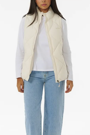 A person is wearing a Rip Curl Anti Series Shorebreak Vest in cream over a white long-sleeved shirt and blue high-waisted jeans. The vest is unzipped, showcasing the shirt beneath. The image features a plain white background.