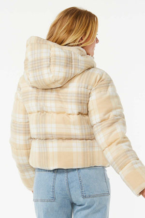 A person with blonde hair, seen from the back, wears a Rip Curl Anti Series Surf Check Puffer with a hood and shower resistance, paired with light blue jeans against a plain white background.