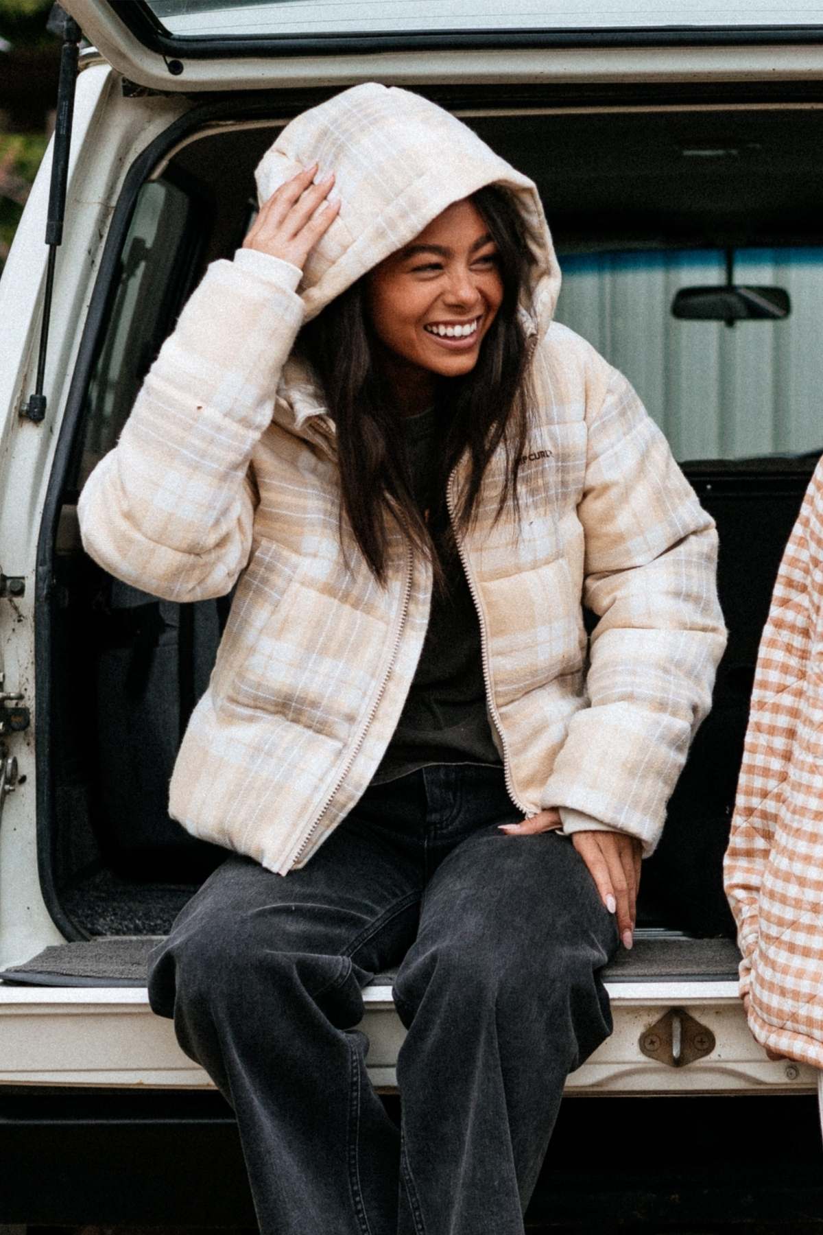 A smiling woman wearing the Rip Curl Anti Series Surf Check Puffer and dark pants sits in the back of a vehicle with the hatch open, touching her hood and looking to the side.