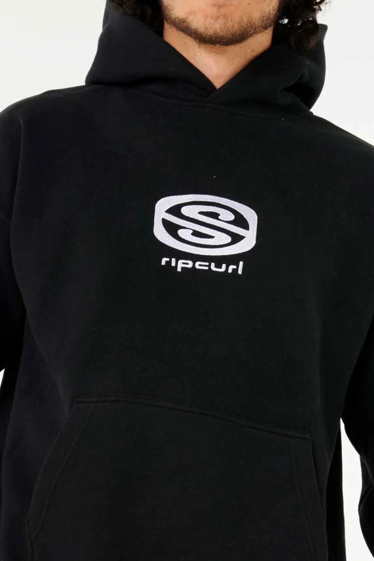 A person wears the Rip Curl Archive Hood, a black, loose-fit hoodie featuring a silver "Rip Curl" logo and text on the chest. Only the lower half of their face and torso are visible, highlighting its classic style.