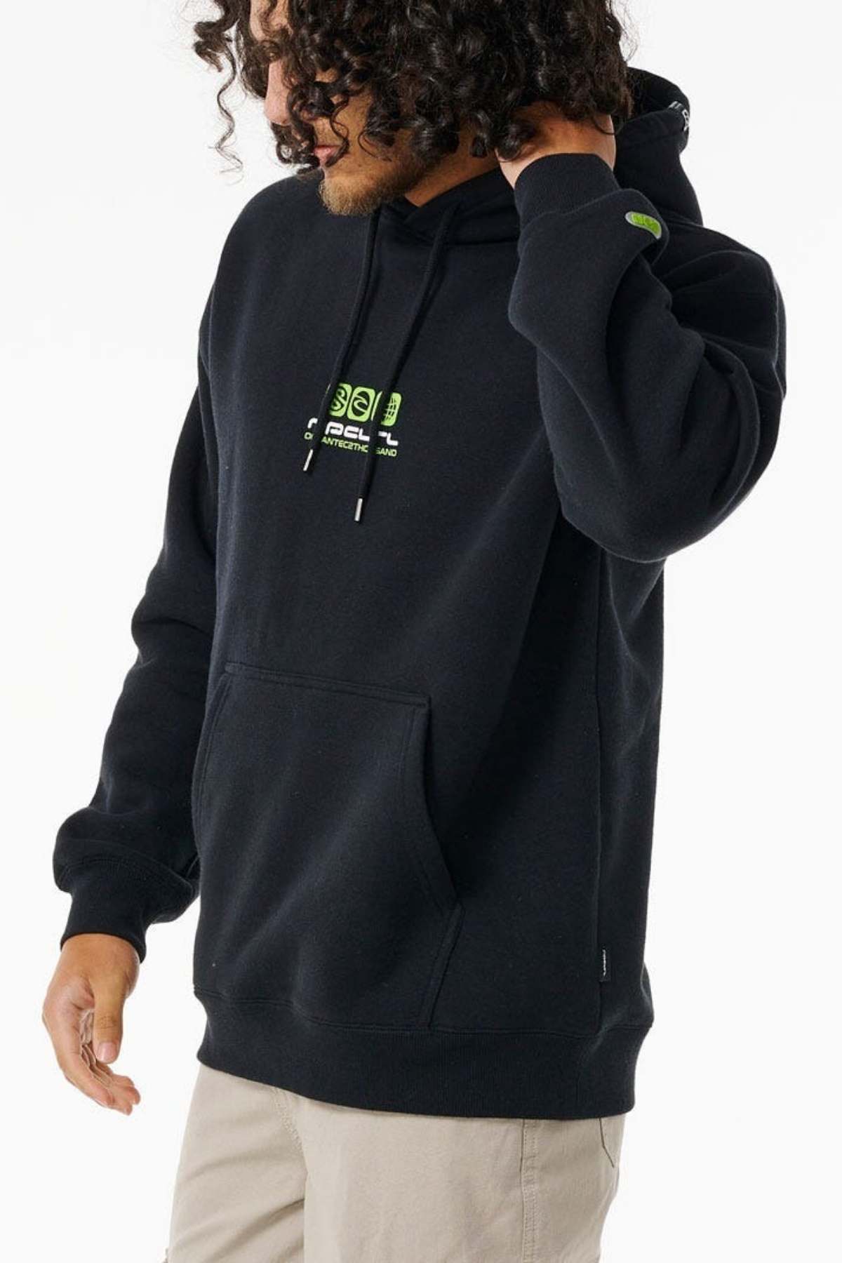 A person with curly hair wears the Rip Curl Archive Ocean Tech Hood in black with neon green text and light beige pants, standing against a white background. They touch their hair while looking down, highlighting surf-inspired style.