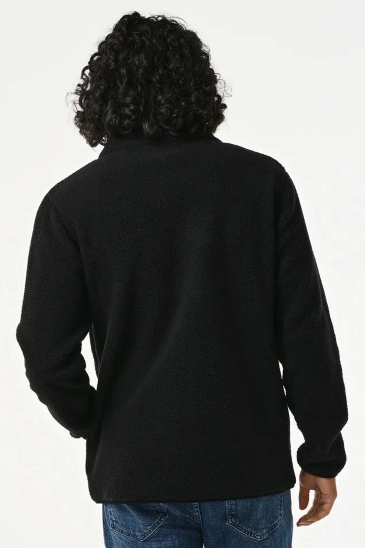 A person with curly hair is seen from the back, wearing a black Rip Curl Bells Polar Fleece jacket and blue jeans against a plain background.