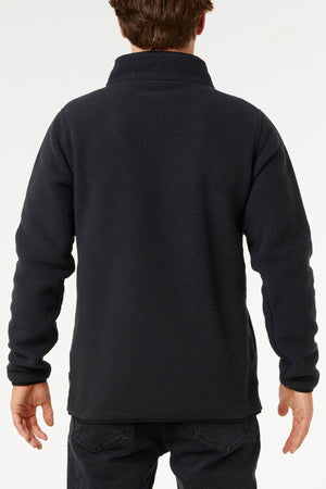 Someone stands facing away from the camera, wearing the Rip Curl Bells Polar Fleece jacket in black with zip pockets and dark jeans, set against a plain light background.