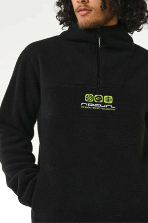A person in a Rip Curl Bells Polar Fleece features a black 1/4 zip with a high collar and neon green logo, perfectly complementing their curly hair against the plain white backdrop.