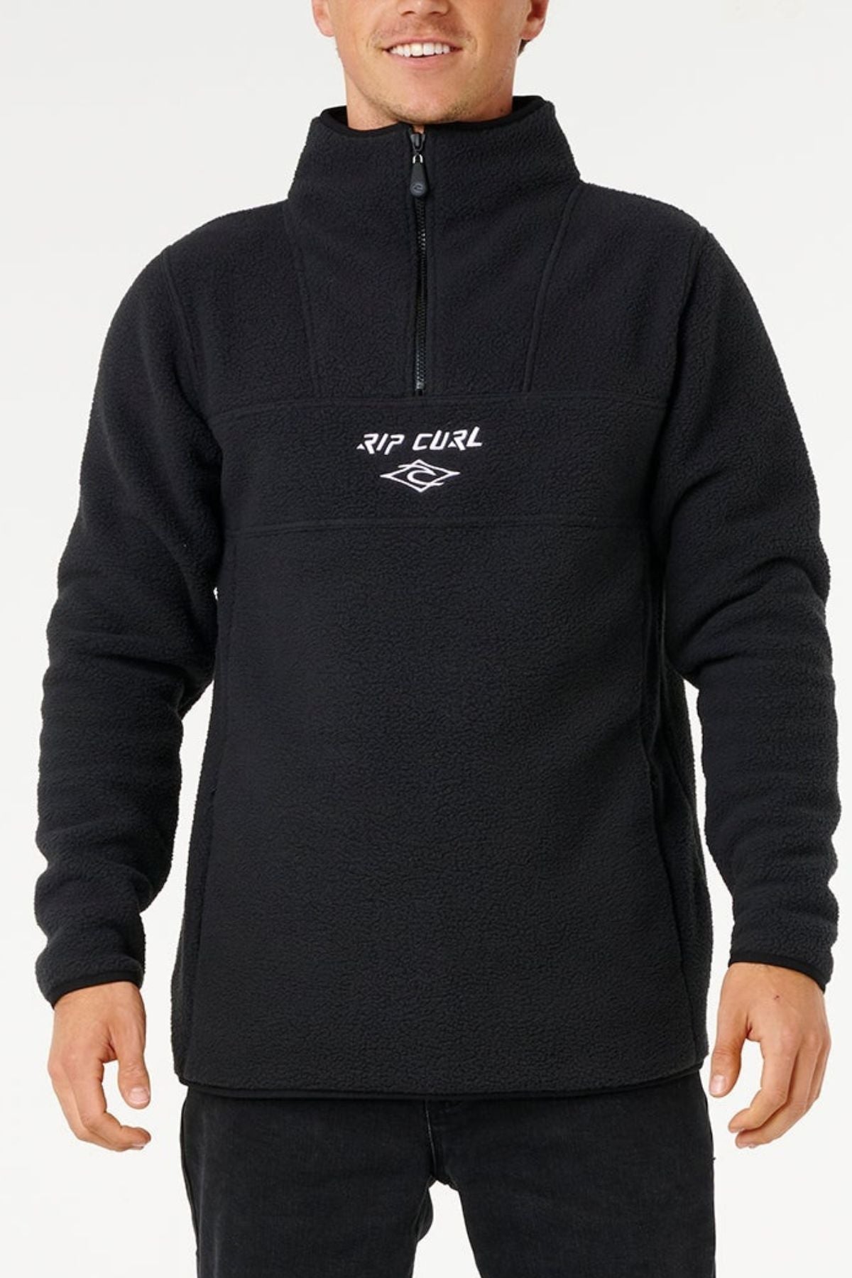 A person in a black Rip Curl Bells Polar Fleece with a front half-zip, zip pockets, and white "RIP CURL" chest embroidery stands against a plain white background.
