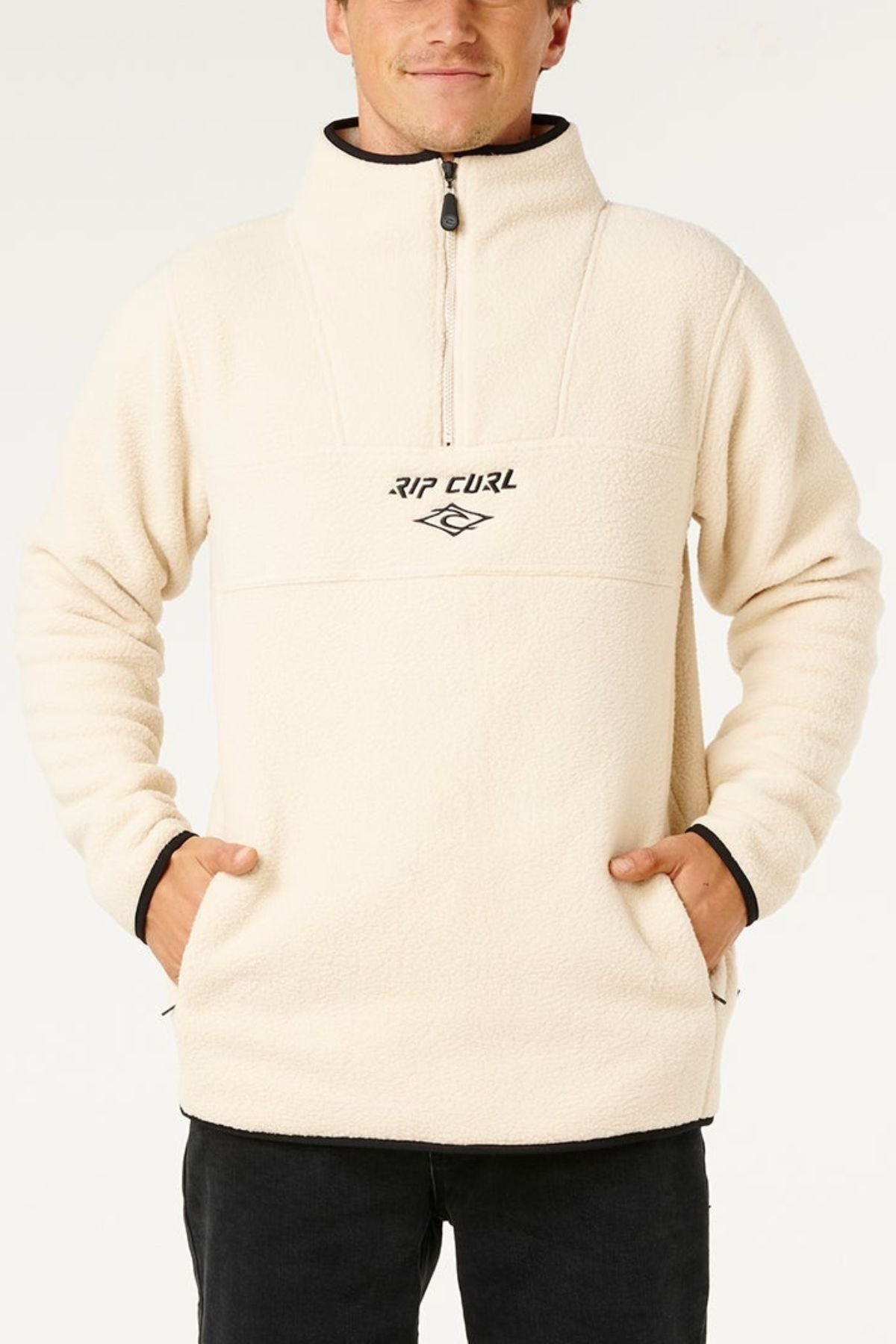 Someone wears a cream Rip Curl Bells Polar Fleece quarter-zip pullover with an embroidered chest logo, standing with hands in pockets against a plain background.