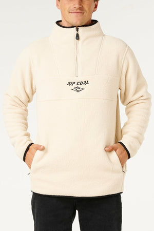 Someone wears a cream Rip Curl Bells Polar Fleece quarter-zip pullover with an embroidered chest logo, standing with hands in pockets against a plain background.