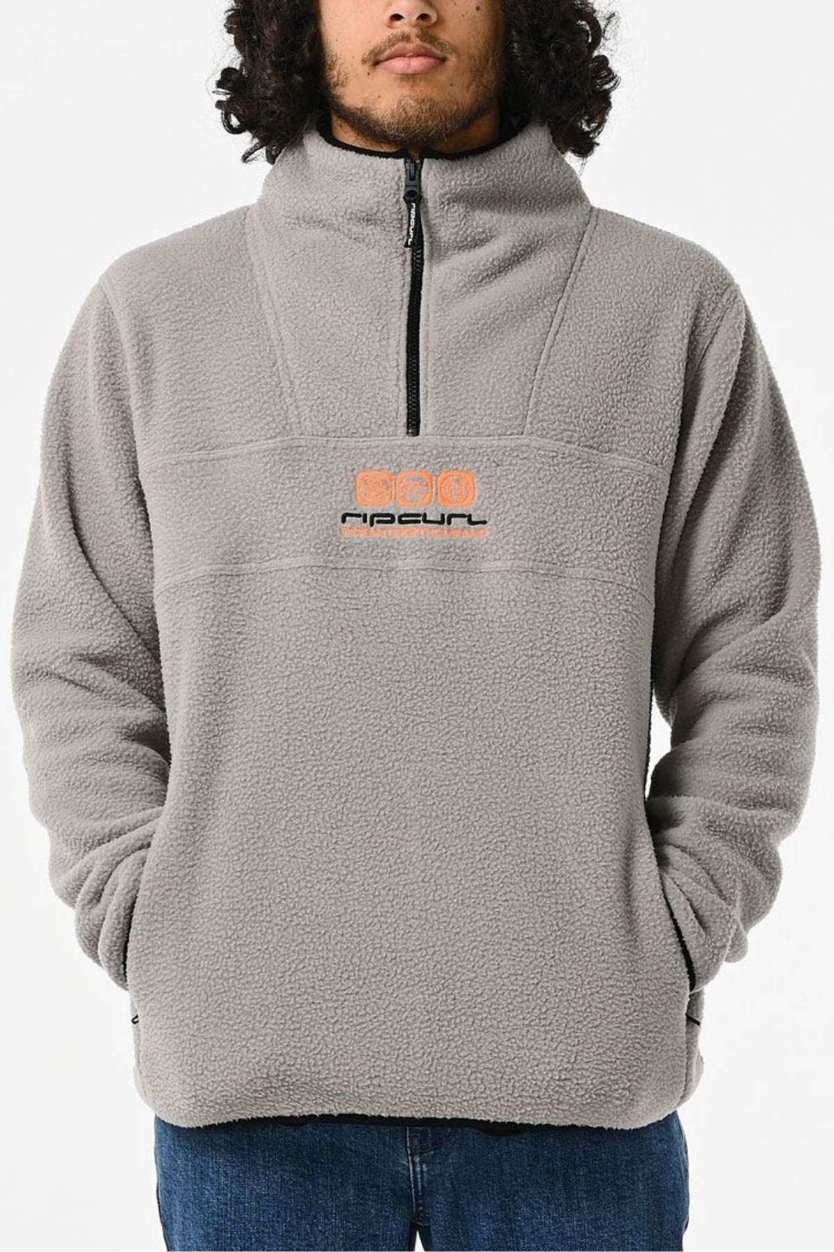 A person sports the Rip Curl Bells Polar Fleece, a gray pullover with a partial zipper and an orange/black logo. Its high collar makes it stand out against the plain white background.