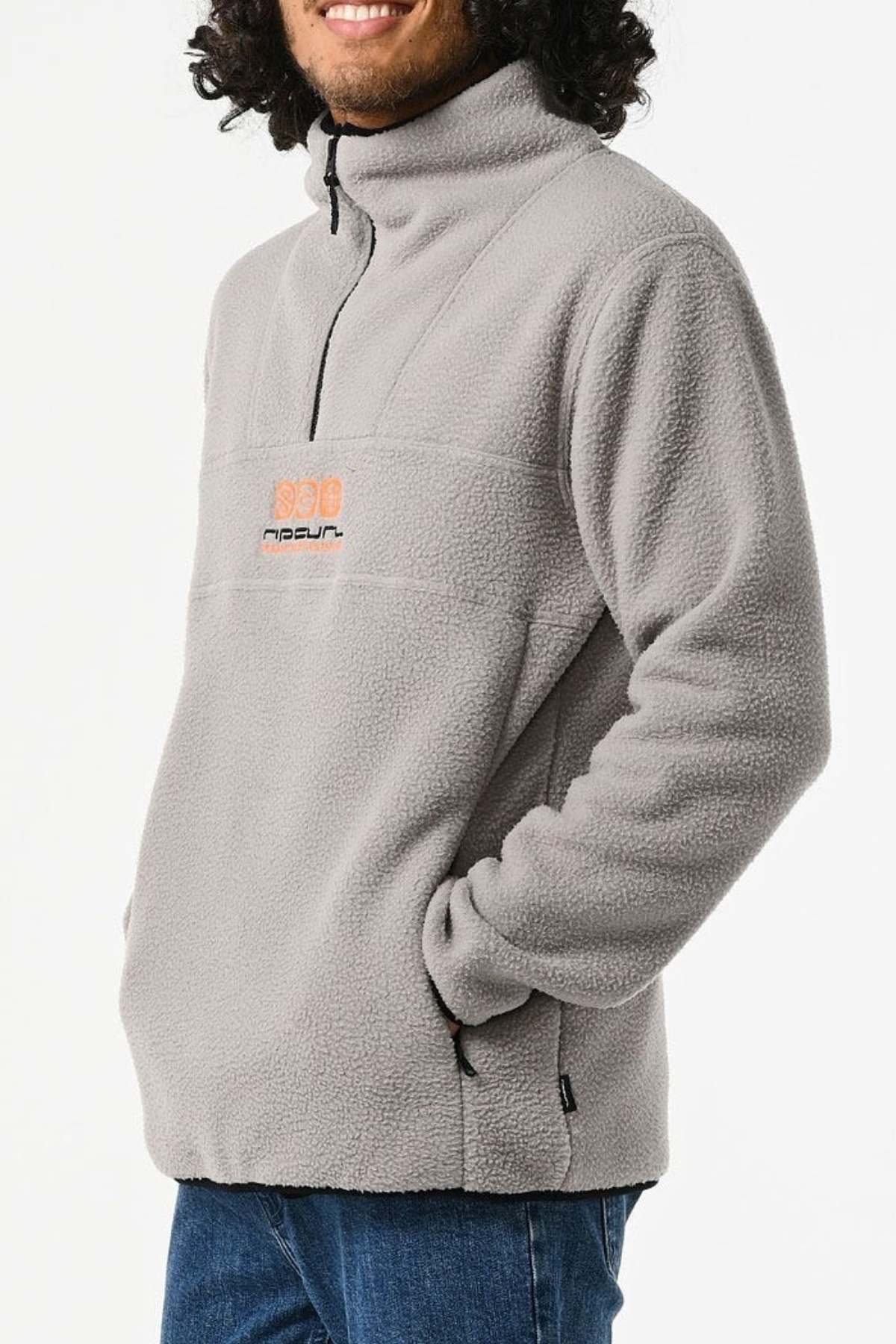 A person is wearing the Rip Curl Bells Polar Fleece, a light gray half-zip pullover with a front pocket. It features a small chest logo. They stand against a white background with arms relaxed in the pockets.