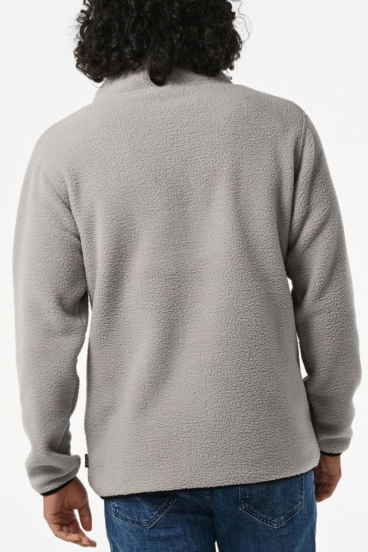 A person with wavy hair stands against a white background, wearing the Rip Curl Bells Polar Fleece in light gray, paired with blue jeans. The jacket exudes a soft, textured polar fleece aesthetic from its rear view.