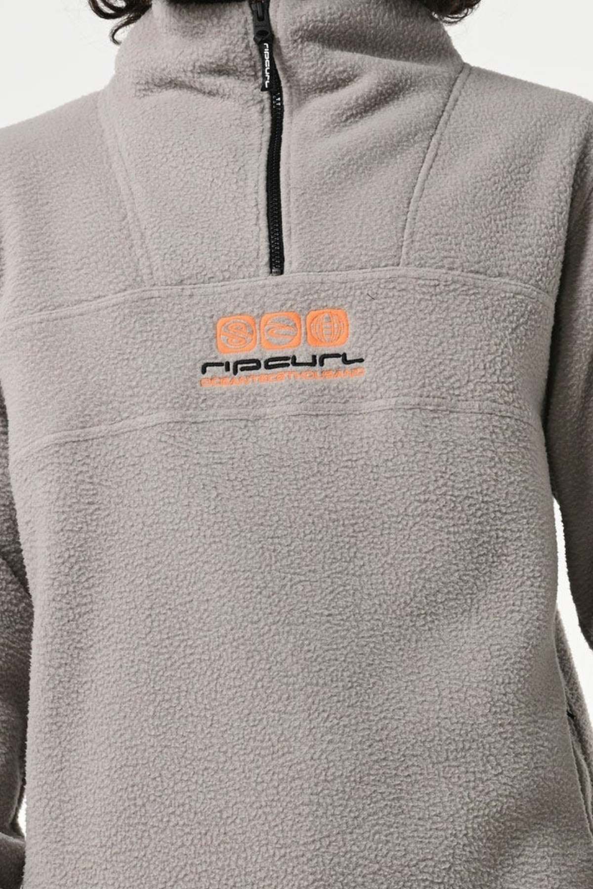 A person wears a Rip Curl Bells Polar Fleece in light gray with a black zipper, featuring an orange and black embroidered "Rip Curl" logo on the chest, showcasing its versatile style and perfect Sherpa material for extra warmth.